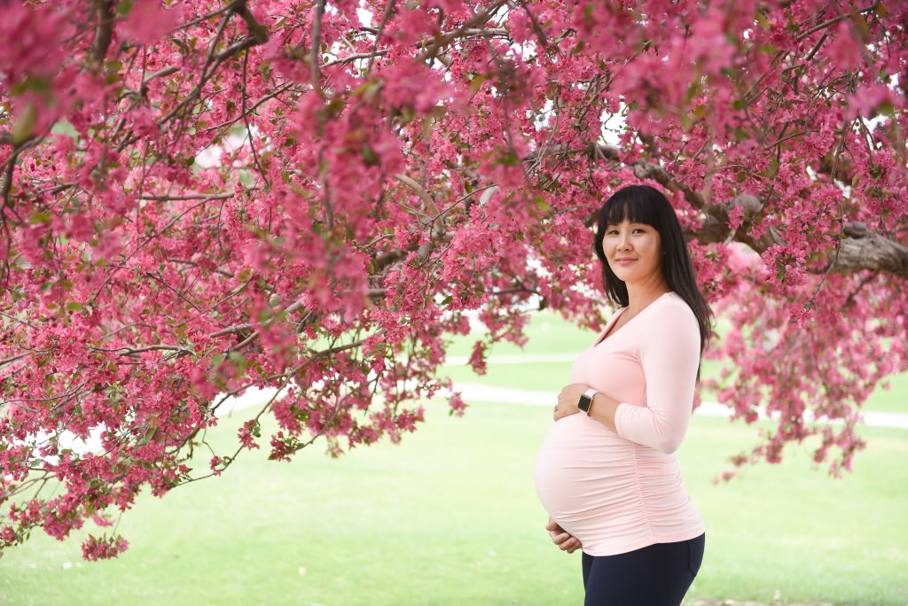 a pregnant women stands under a pink blooming tree