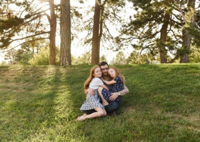 a father sits on a grassy hill while he is photographed with his two daugthers