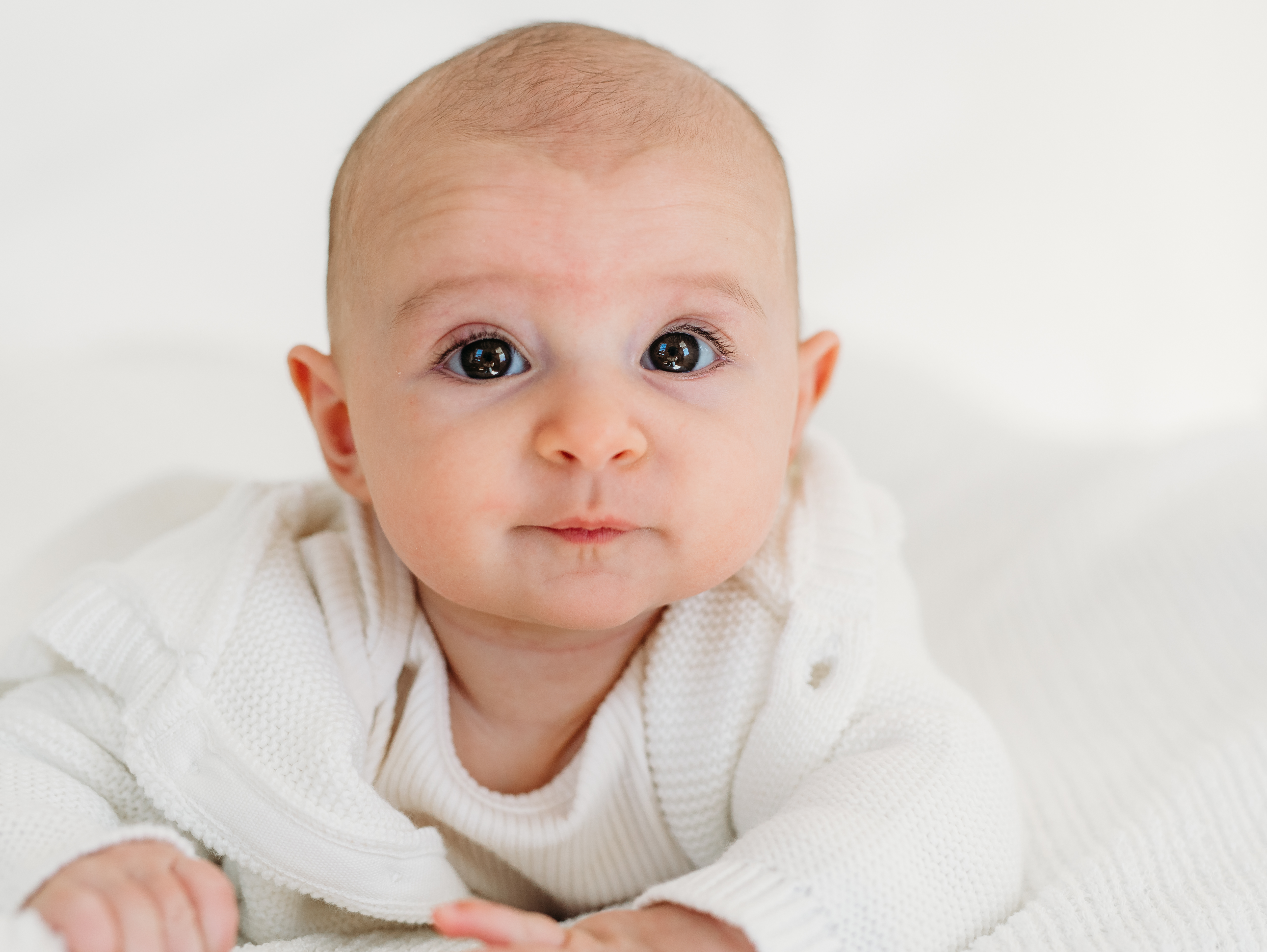 a 3 month old baby girl on her belly looks at the camera 